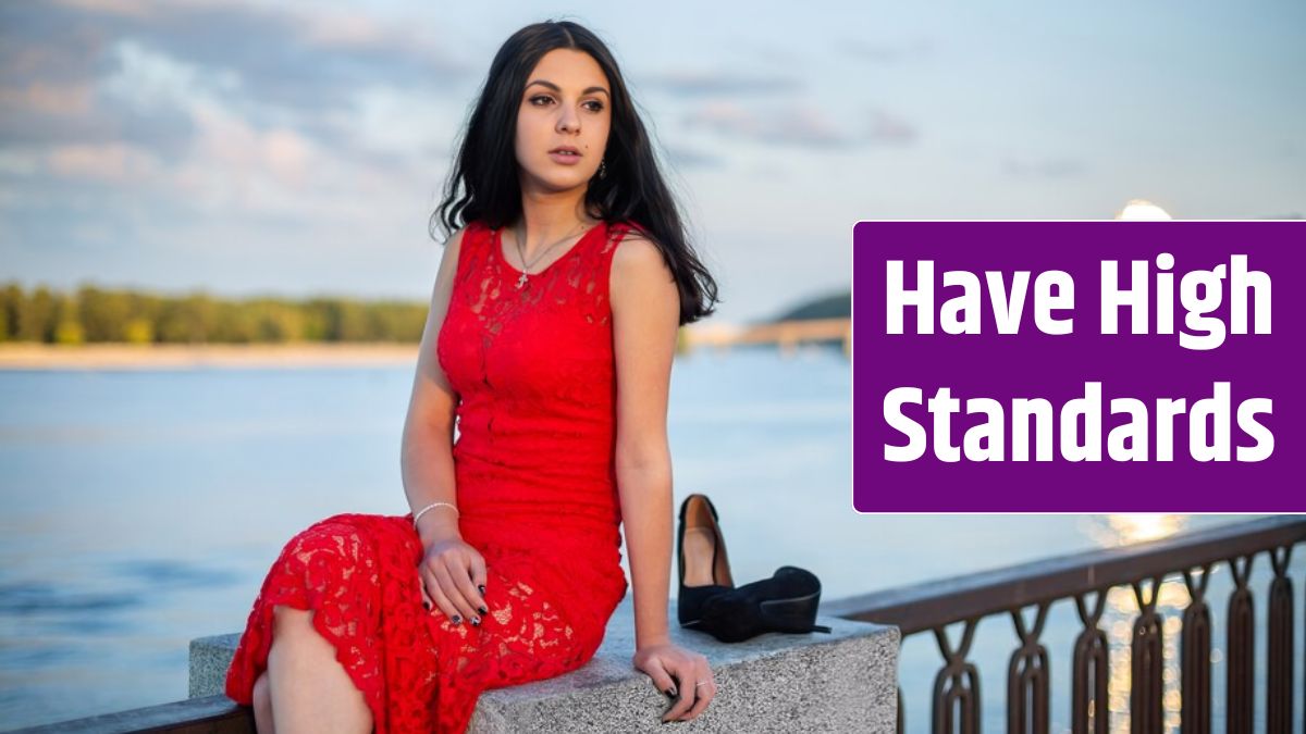 A girl in a long red dress is sitting on the fence of the river embankment.